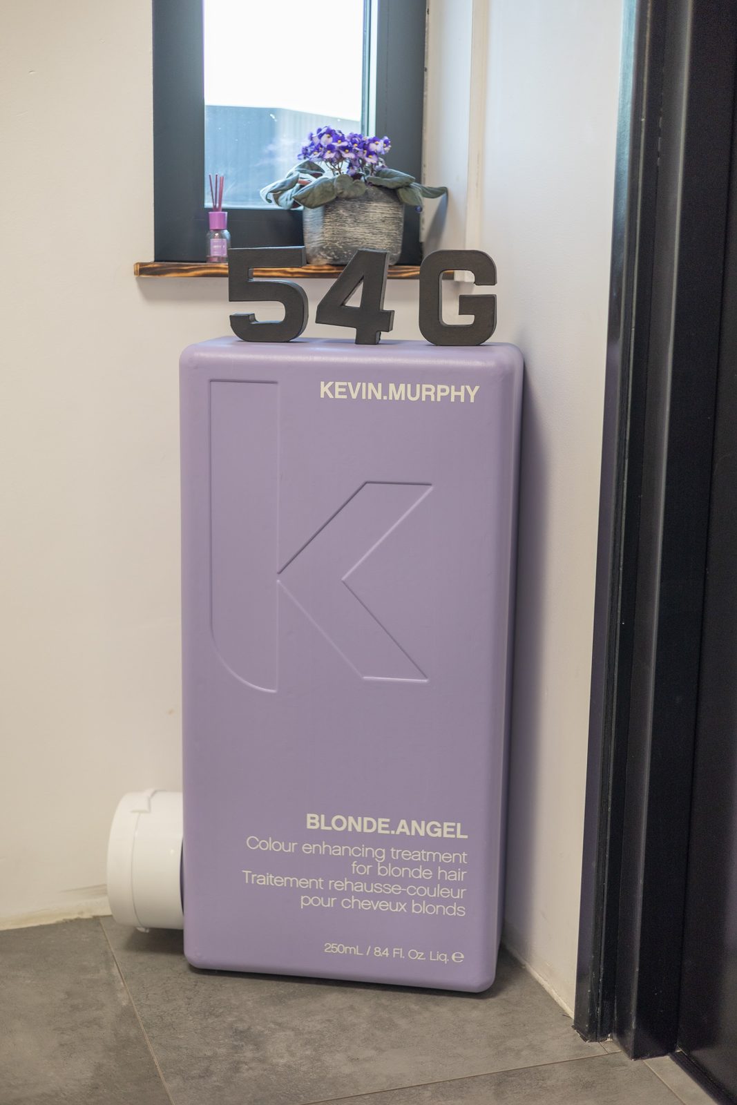Een grote lila fles KEVIN.MURPHY BLONDE.ANGEL tegen de muur